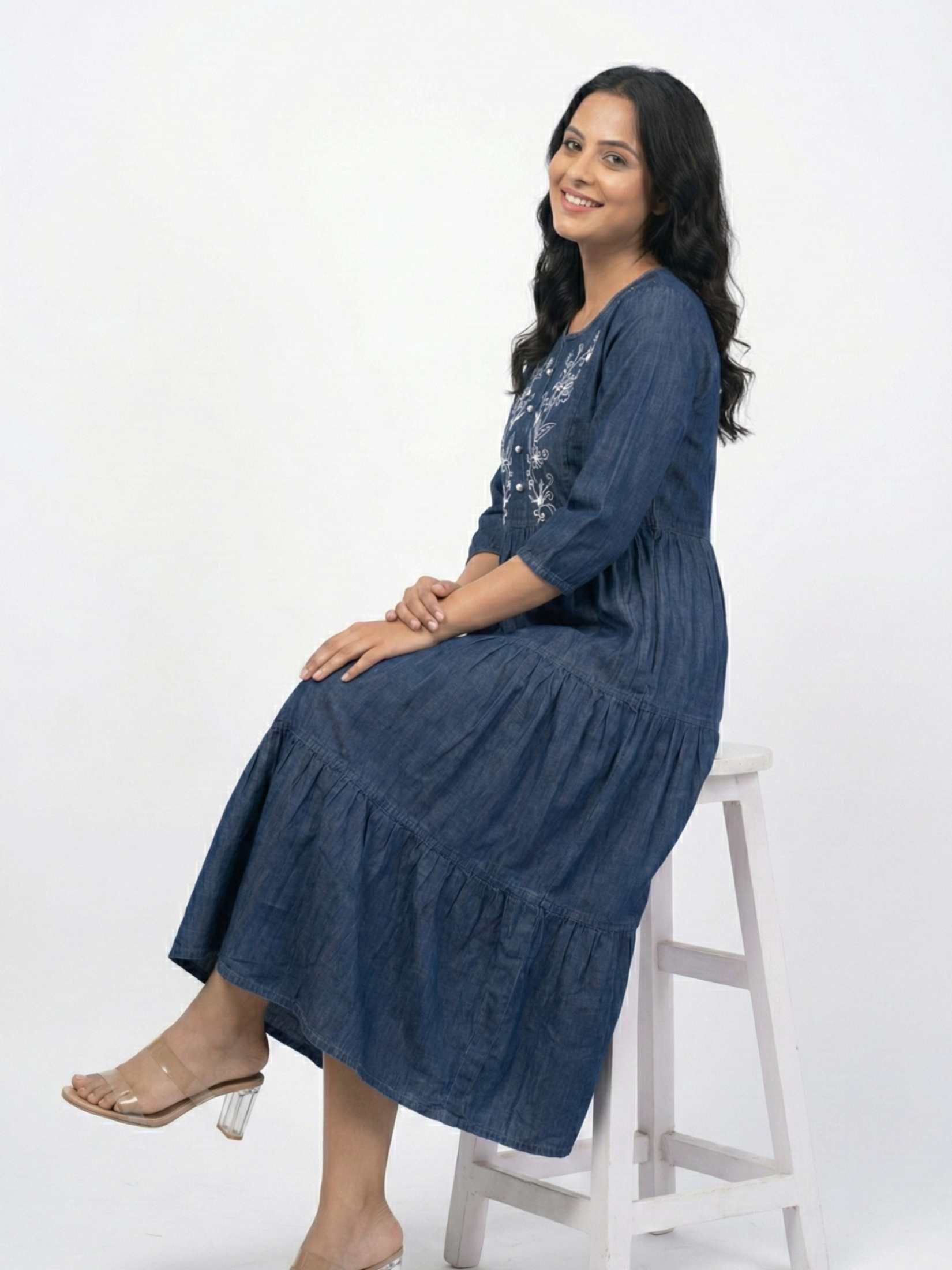 Woman wearing a blue dress sitting on a white stool against a white background

