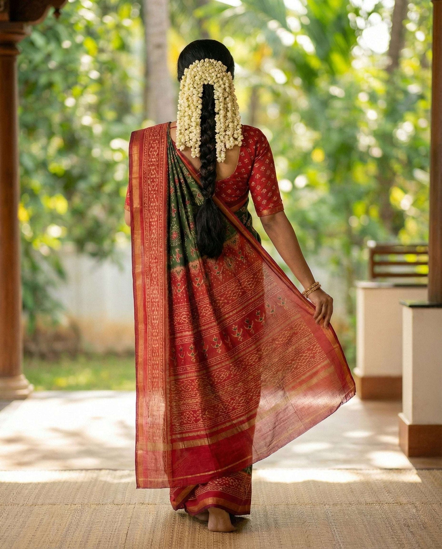 A beautiful model in a traditional Kerala setting wearing a Jodhpuri cotton saree. The saree features a deep forest green body with intricate cream and red tribal patterns, a broad crimson red border with golden zari lining, and a matching red blouse with a gold diamond print back