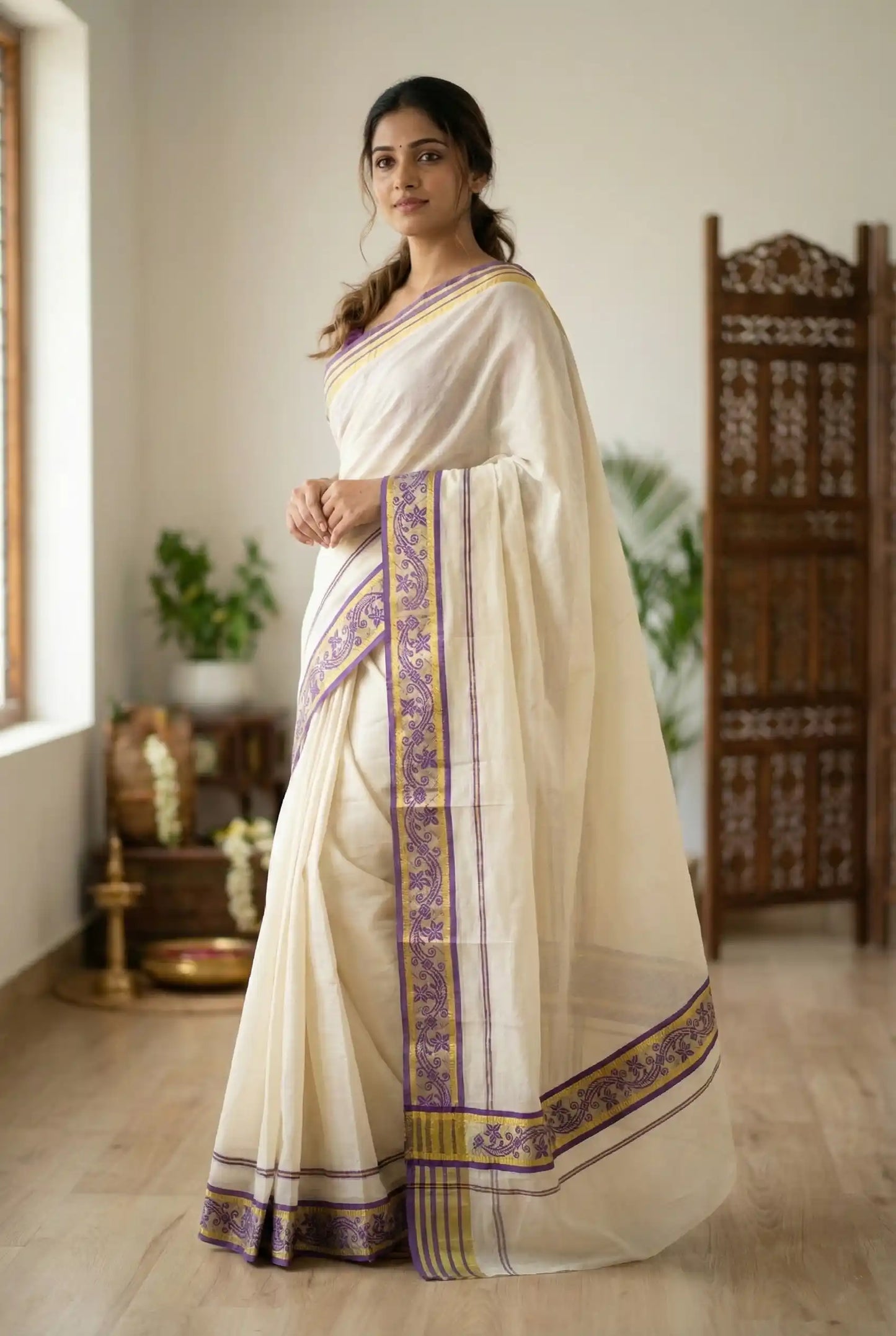 Woman wearing a traditional white saree with purple and gold border in a room.

